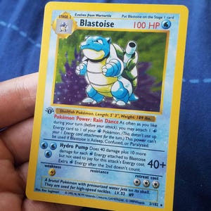 May include: A 1999 Blastoise trading card featuring a blue, turtle-like creature with cannons on its shell. The card displays 100 HP, the Rain Dance power, and the Hydro Pump attack. It includes text details like Shellfish Pokémon, 5' 3" length, and 189 lbs. weight.