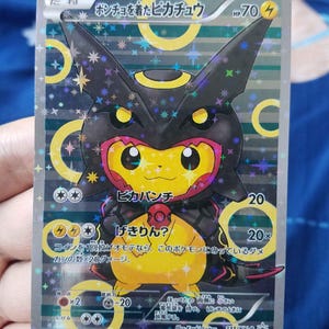 May include: A holographic trading card featuring a yellow Pikachu character wearing a black Umbreon-themed poncho. The card displays Japanese text, an HP of 70, and a lightning energy symbol. The background has yellow rings and a star-patterned foil finish.