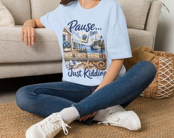 May include: A light blue t-shirt featuring a graphic print of a coastal scene with a lighthouse, sailboats, and mahjong tiles. The text reads "Pause... Oops" at the top and "Just Kidding" at the bottom, with a starfish and rope accent.