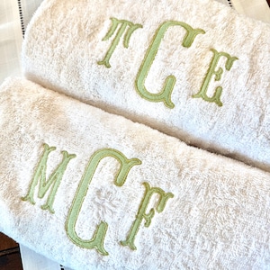 May include: Two folded white terry cloth towels featuring embroidered monograms in a light green thread. The top towel displays the letters TCE, while the bottom towel features the letters MCF, both stitched in a decorative serif font style.