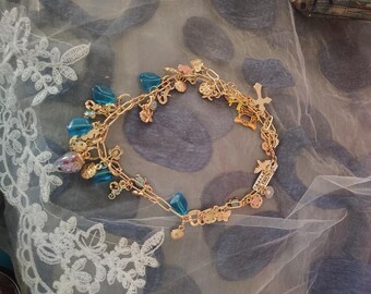 Può includere: Un braccialetto con ciondoli color oro caratterizzato da una varietà di pendenti, tra cui perline blu traslucide, piccole forme metalliche e un ciondolo rettangolare con testo, collegati da una sottile catena a maglie.