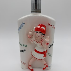 May include: A white ceramic decanter with a silver-colored metal stopper. The front features a raised, three-dimensional figure of a person in a red hat and white dress. The surface is decorated with various text including TOOTSIE and Hot Lips.