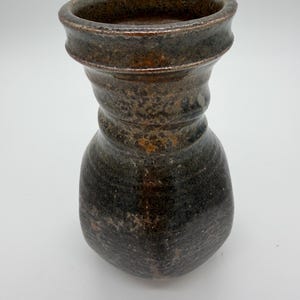 May include: A ceramic vase with a dark brown and black mottled glaze. The vessel features a wide, flared rim that tapers into a narrow neck, expanding into a rounded, bulbous body. The surface has a textured, uneven finish with visible variations in color.
