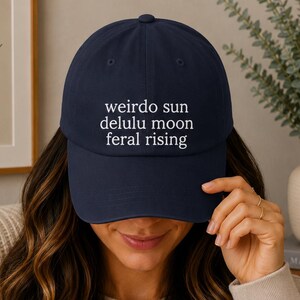 May include: A navy blue baseball cap featuring white embroidered text on the front that reads: weirdo sun, delulu moon, feral rising. The cap has a structured crown, a curved brim, and a button on top, constructed from a solid fabric material.