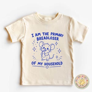 May include: A cream-colored short-sleeve t-shirt featuring a blue graphic of a mouse holding a bottle. The text reads I AM THE PRIMARY BREADLOSER OF MY HOUSEHOLD. The design includes small star accents and a heart symbol below the mouse illustration.