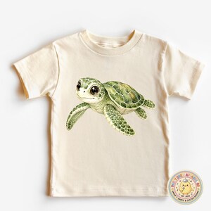May include: A cream-coloured short-sleeve t-shirt featuring a watercolour-style illustration of a green sea turtle with large dark eyes. In the bottom right corner, there is a circular logo with a yellow chick and the text Mudy Chew Apparel Co.