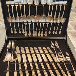 May include: A silver-toned flatware set arranged in a black storage case. The collection includes various forks, spoons, and knives with ornate handles. The pieces are organized in two rows, with the upper section holding spoons and forks, and the lower section containing knives and additional forks.