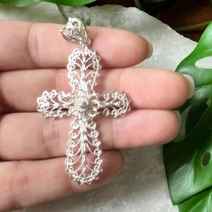 May include: A silver filigree cross pendant with a floral design. The pendant is large and intricate, with a delicate, openwork design.
