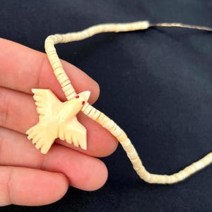 May include: Ivory-colored bird pendant necklace with red accents on the eyes. The bird is carved with detailed wings and a pointed beak. The necklace features a strand of small, off-white shell beads.