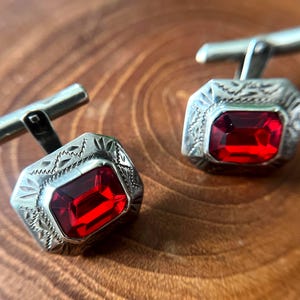 May include: A pair of silver-toned metal cufflinks featuring a rectangular, faceted red stone set in the center. The metal frame surrounding the stone has an engraved, textured pattern along the edges and a hinged T-bar closure on the back.