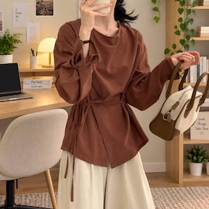 Op de afbeelding: Een bruine blouse met lange mouwen, een overslag aan de voorzijde met een striksluiting in de taille, gecombineerd met een crèmekleurige wijde broek en een tweekleurige bruin-crème handtas met twee hengsels.