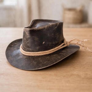 May include: A dark brown, distressed-finish hat with a pinched crown and a wide, slightly curved brim. A thick, light tan braided rope band encircles the base of the crown, featuring a knotted detail with loose ends extending from the side.