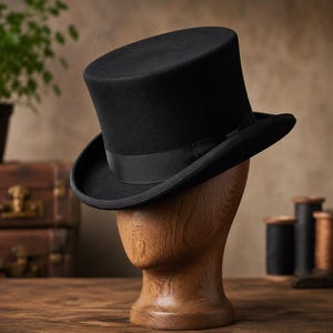 Puede incluir: Un sombrero de copa negro clásico con corona plana y ala curva, que presenta una cinta de grosgrain negra a juego alrededor de la base. El sombrero se exhibe sobre una cabeza de maniquí de madera clara y lisa, mostrando su forma.