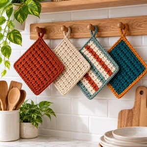 May include: Four square crochet potholders hang from a wooden rack. The first is solid rust, the second is cream, the third features cream, rust, and teal stripes, and the fourth is teal with a mustard yellow border. Each has a small hanging loop.