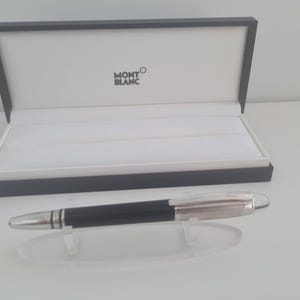 Peut inclure: Un stylo à bille noir et argenté avec un clip métallique et une prise texturée, posé sur un support en acrylique transparent devant une boîte de présentation ouverte noire et blanche avec le logo Montblanc imprimé sur le couvercle intérieur.
