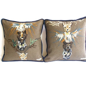 Puede incluir: Dos cojines de sofá marrones con ribete azul marino presentan diseños de animales bordados. Cada cojín representa un tótem de animales, incluyendo un ciervo, un búho, un oso y un lobo. Los diseños están en tonos de marrón, naranja, amarillo, verde y azul.