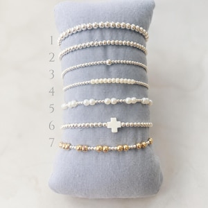 Op de afbeelding: Zeven elastische armbanden genummerd van één tot zeven. Armband één heeft kleine zilveren kralen. Twee heeft middelgrote zilveren kralen. Drie heeft kleine zilveren kralen met één grotere kraal. Vier heeft kleine witte parels. Vijf heeft grotere witte parels. Zes heeft een wit kruisje. Zeven heeft afwisselend gouden en zilveren kralen.