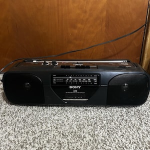 Könnte beinhalten: Ein schwarzer tragbarer Sony Radio-Kassettenrekorder mit zwei seitlichen Lautsprechern, einer zentralen analogen Radioskala für FM- und AM-Frequenzen, einem Kassettenfach und einer ausziehbaren Antenne an der Oberseite des Geräts.