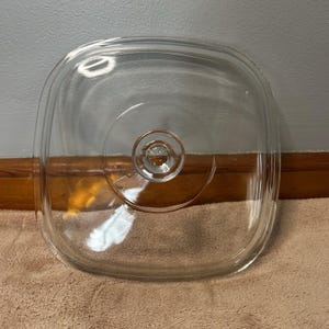 Puede incluir: Una tapa de vidrio transparente cuadrada con esquinas redondeadas y un pomo central circular. La tapa presenta una superficie lisa y transparente con un borde elevado en el perímetro y una sección circular hundida alrededor del pomo central.