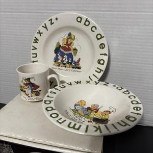 以下が含まれることがあります： プレート、ボウル、マグカップの3点セットの子供用陶磁器食器セット。各アイテムはクリーム色で、緑色のアルファベットの縁取りが施されています。プレートにはメアリー・メアリー、マグカップには騎手、ボウルには桶に入った3人の男性が描かれています。