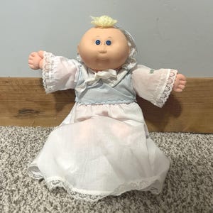 Könnte beinhalten: Eine Stoffpuppe mit rundem Kopf, blauen Augen und einem kleinen Büschel gelber Wollhaare. Die Puppe trägt ein weißes Kleid mit Spitzenbesatz an den Ärmeln und am Saum, kombiniert mit einer hellblauen, karierten Weste mit weißer Schleife.