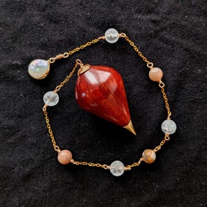 May include: A teardrop-shaped red wooden pendulum with a gold-toned metal tip, attached to a delicate gold-toned chain. The chain is accented with small, round, translucent and opaque beads in shades of white, peach, and light brown.
