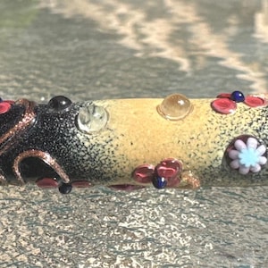 May include: A cylindrical glass bead featuring a gradient from black to a central beige section. The surface is decorated with copper-colored swirling lines, small pink and blue floral motifs, and tiny clear and dark spherical glass accents.