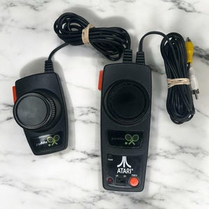 May include: A pair of black Atari paddle controllers with circular knobs, orange side buttons, and black cables. The main unit features the Atari logo, a reset button, an on-off-menu switch, and a small red indicator light. The controllers include yellow and white RCA plugs.