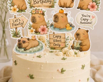 May include: A round, off-white frosted cake decorated with small green leaves and white pearls, topped with multiple capybara-themed paper cutouts featuring text like Happy Birthday, Let's Pawty, Capy Vibes Only, and You're Capy-Awesome.