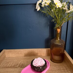 May include: A bright pink oval tray holding a small black bowl with a light pink rose-shaped object inside, positioned next to a ribbed amber glass vase filled with white daisy-like flowers with yellow centres, set against a dark blue wall.