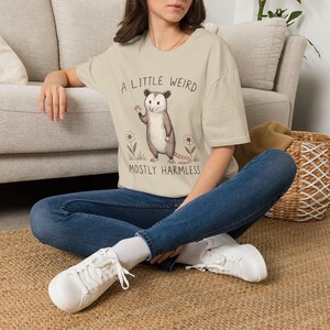 Peut inclure: Un t-shirt beige à manches courtes orné d'une illustration graphique d'un opossum faisant un signe de la main. Le texte A LITTLE WEIRD et MOSTLY HARMLESS est imprimé au-dessus et en dessous de l'animal, entouré de deux petites fleurs blanches.