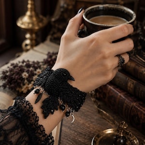 May include: A black crocheted bird bracelet featuring a detailed raven design with dangling silver-toned charms, including a small crescent moon, black beads, and leaf-shaped accents, secured around the wrist with a textured black crochet band.