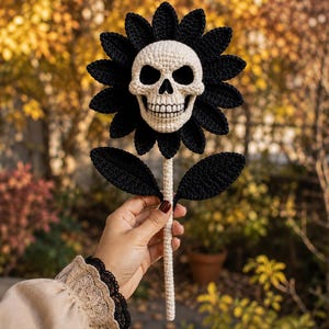 May include: A handcrafted crochet flower featuring a central off-white skull with detailed eye sockets and teeth, surrounded by a ring of pointed black petals. The flower is attached to a long, slender off-white stem with two black leaves.