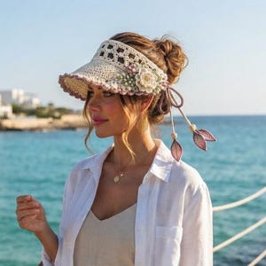 May include: A beige crochet visor with a floral accent and dangling leaf-shaped charms, paired with a beige sleeveless top, beige shorts, and a white button-down shirt. A woven straw tote bag is carried alongside the outfit near the ocean.