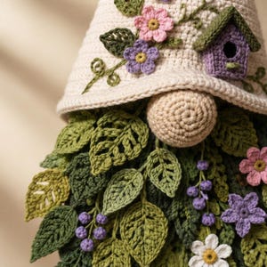 Op de afbeelding: Een gehaakte kabouter met een cr&egrave;mekleurige muts versierd met roze en paarse bloemen en een klein paars vogelhuisje. De kabouter heeft een ronde beige neus en een dichte baard bestaande uit diverse groene gehaakte bladeren, kleine paarse bessen en witte, roze en paarse bloemen.
