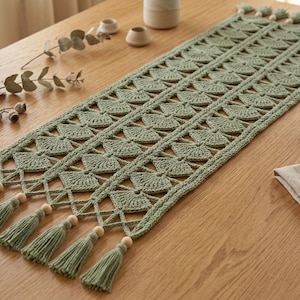 May include: A sage green rectangular crochet table runner featuring a repeating fan-like stitch pattern, finished with light wooden beads and matching sage green tassels at both ends, creating a textured and intricate textile design.