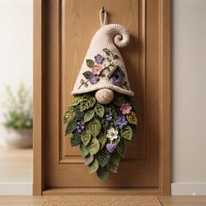 Op de afbeelding: Een gehaakte kabouter als wandhanger met een cr&egrave;mekleurige puntmuts met een gekrulde punt, versierd met kleine paarse en roze bloemen en een vogelhuisje. De baard bestaat uit gelaagde groene bladeren met paarse bessen en bloemen.