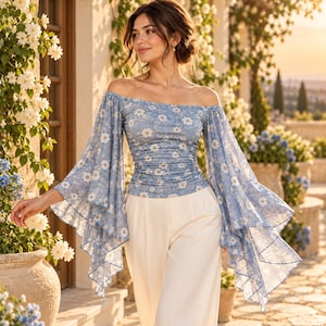May include: An off-the-shoulder top in a light blue fabric featuring a white daisy floral pattern. The garment has long, wide, flared sleeves with a ruffled hem and a fitted, ruched bodice. It is paired with plain white trousers.