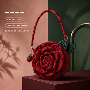 May include: A round, red crocheted handbag shaped like a blooming flower with layered petals, a single braided handle, and two dark green crocheted leaves. A small red strawberry charm hangs from the handle. Text reads PDF PATTERN INSTANT DOWNLOAD INTERMEDIATE LEVEL.
