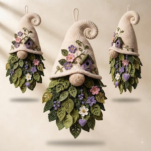 Op de afbeelding: Drie hangende gehaakte kabouterornamenten met cr&egrave;mekleurige puntmutsen met gekrulde uiteinden. Elke muts is versierd met kleine paarse vogelhuisjes, roze en paarse bloemen en groene ranken. De baarden zijn gemaakt van gelaagde groene gehaakte bladeren.