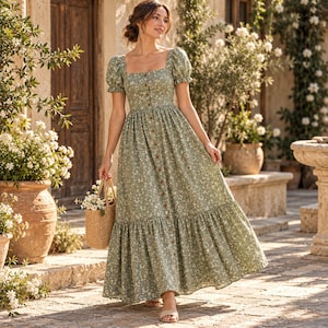 Può includere: Un abito midi verde salvia con scollo quadrato, maniche corte a sbuffo e una gonna lunga con orlo a balze. L'abito è decorato con una piccola stampa floreale bianca e una fila verticale di bottoni marroni che corre lungo il centro anteriore.