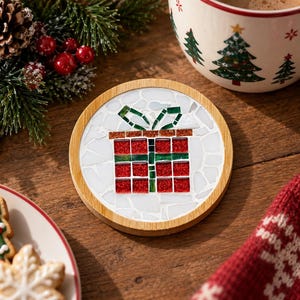 Puede incluir: Un posavasos circular con marco de madera clara que presenta un diseño de mosaico de un regalo rojo con un lazo verde. El fondo del mosaico está hecho de piezas blancas de formas irregulares, mientras que el regalo utiliza azulejos rojos y verdes.