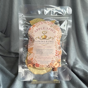 May include: A clear plastic pouch containing a simmer pot blend of dried apple slices, cinnamon sticks, and botanicals. The label features the text Apple Spice, Simmering Intentions, and Simmer Pot Blend, surrounded by a decorative autumn leaf border.