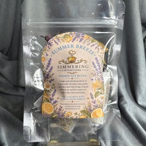 May include: A clear plastic pouch containing a simmer pot blend labeled Summer Breeze. The label features illustrations of lavender, honeysuckle, and lemon slices, with text describing a blend of honeysuckle, lavender, chamomile, and lemon peel.