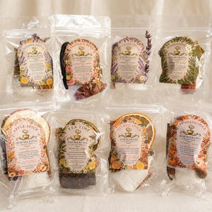 May include: Eight clear plastic pouches containing Simmering Intentions stovetop potpourri blends. Each pouch features a decorative label with the scent name, including Summer Breeze, Vanilla Rose, Lavender Dream, Scarborough Fair, Apple Spice, Tea Time, Citrus Spice, and Pumpkin Spice, alongside dried botanicals, fruit slices, and spices.