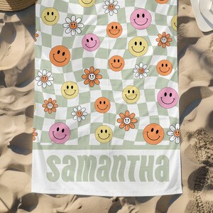May include: A white beach towel with a green and white checkered pattern. The towel has a repeating pattern of smiley faces and flowers in pink, yellow, and orange. The text "Samantha" is printed in green at the bottom of the towel.
