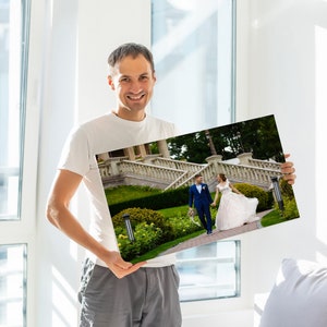 Op de afbeelding: Een man houdt een gedrukte foto vast van een bruidspaar dat een trap afloopt buiten een gebouw. De foto is in kleur en toont het paar in formele kleding.