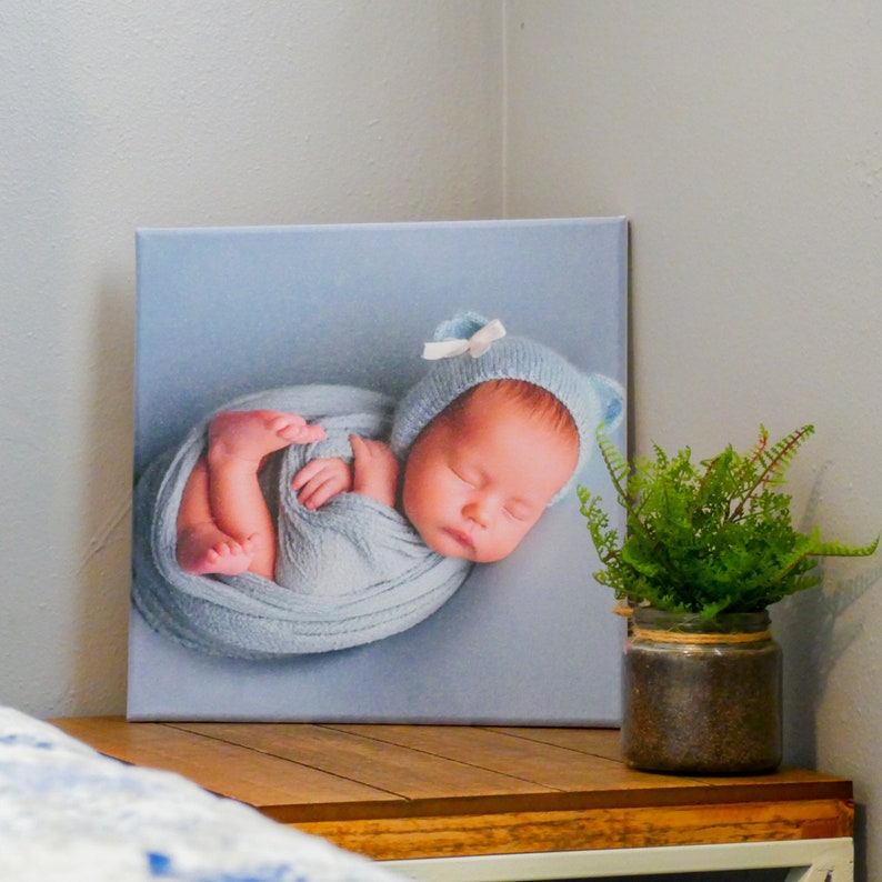 Op de afbeelding: Een canvas print van een slapende baby gewikkeld in een blauwe deken met een witte strik op zijn muts.
