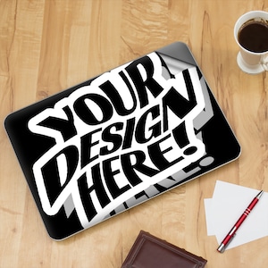 Op de afbeelding: Een zwarte laptop met een witte sticker waarop "YOUR DESIGN HERE!" staat. De sticker is gedeeltelijk losgekomen, waardoor de zwarte oppervlakte van de laptop zichtbaar wordt.