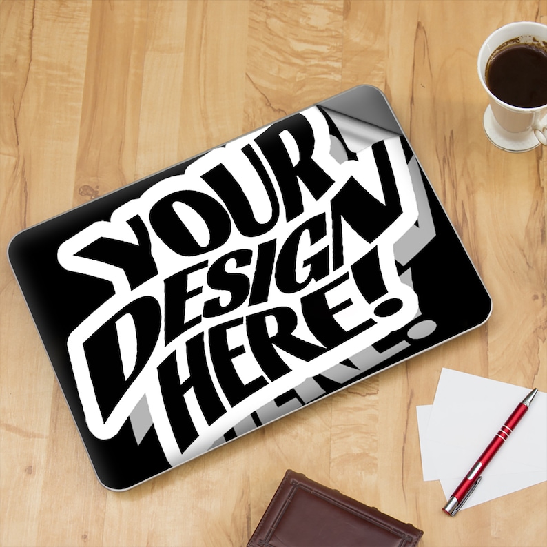 Op de afbeelding: Een zwarte laptop met een witte sticker waarop "YOUR DESIGN HERE!" staat. De sticker is gedeeltelijk losgekomen, waardoor de zwarte oppervlakte van de laptop zichtbaar wordt.